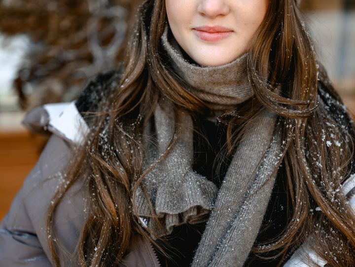 a girl in a brown jacket and hat stands outside and smiles. there is snow on her hair. winter