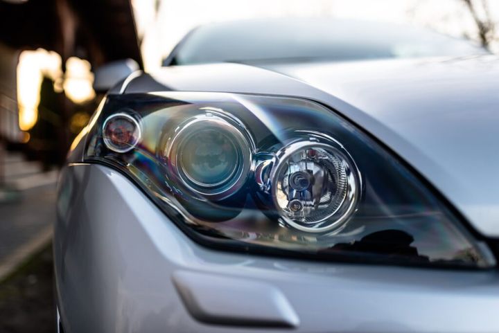 Front headlight of a silver, modern car with three lamps in the middle, visible headlight washer.