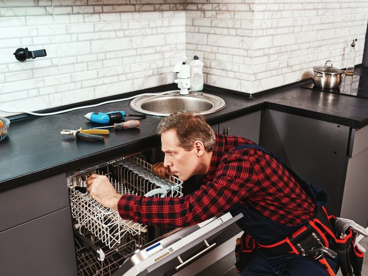 Side view of repairing dishwasher. Male technician sitting near dishwasher