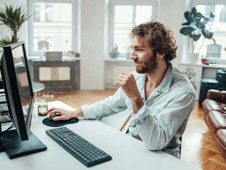 Joyful guy with curly hairs and beard sits at table working on pc