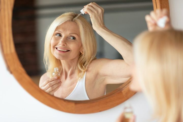 Happy mature woman applying serum or natural oil on her scalp, looking at her reflection in mirror