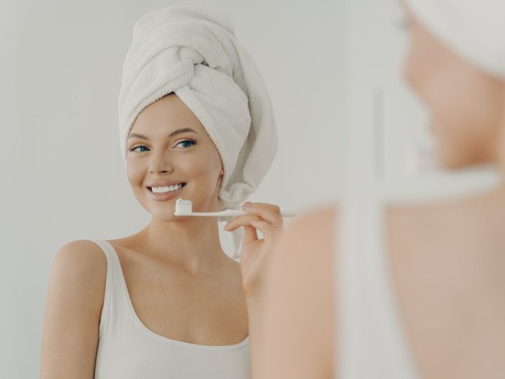 Beautiful young woman with healthy perfect smile brushing teeth and looking in mirror