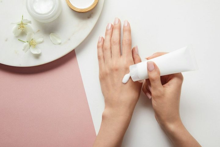 cropped view of woman applying cream, holding hands near plate with cosmetics and flowers on white