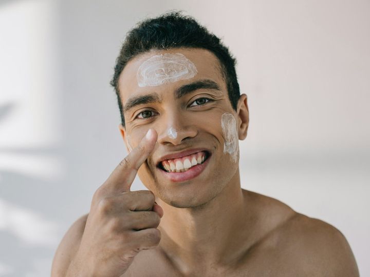 handsome mixed race man applying cosmetic cream on face with finger and smiling while looking away