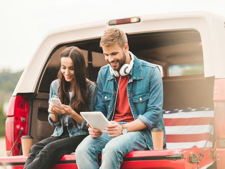 happy young couple using gadgets while sitting in car trunk during trip