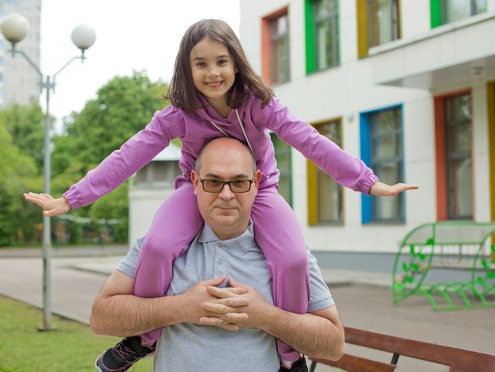 A cute daughter in a purple tracksuit sits on her father's neck, in spring, in the city