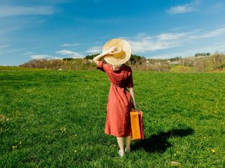 Beautiful girl in red dress and hat with suitcase on meadow