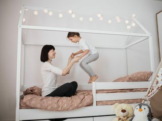 Cheerful mum babysitter play with cute active small kid girl jump on bed