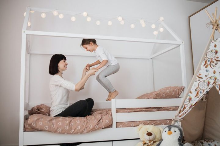 Cheerful mum babysitter play with cute active small kid girl jump on bed