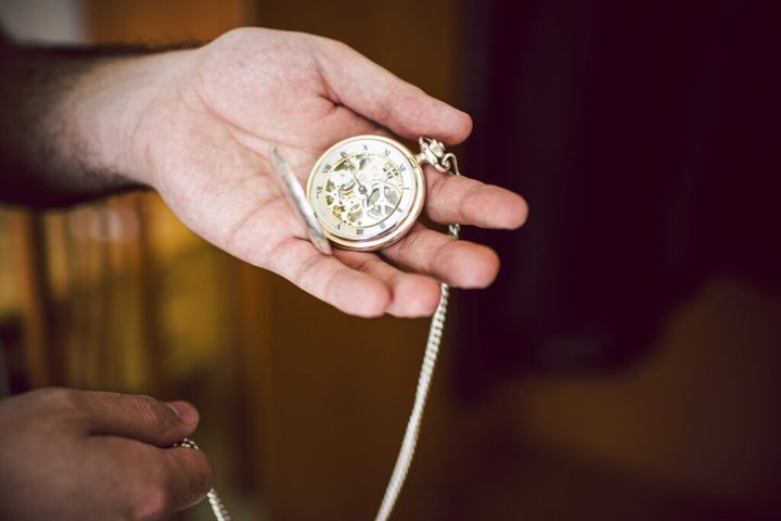 A man's hand holds an antique pocket watch with its gears and hand in sight.