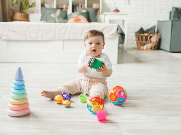 baby 6-9 months old playing with a colorful rainbow toy pyramid sitting in a white sunny bedroom.
