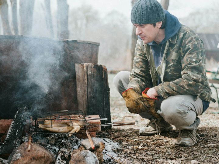 young man survivalist cooks roasts chicken meat food are fried on grill on smoldering coals or ember